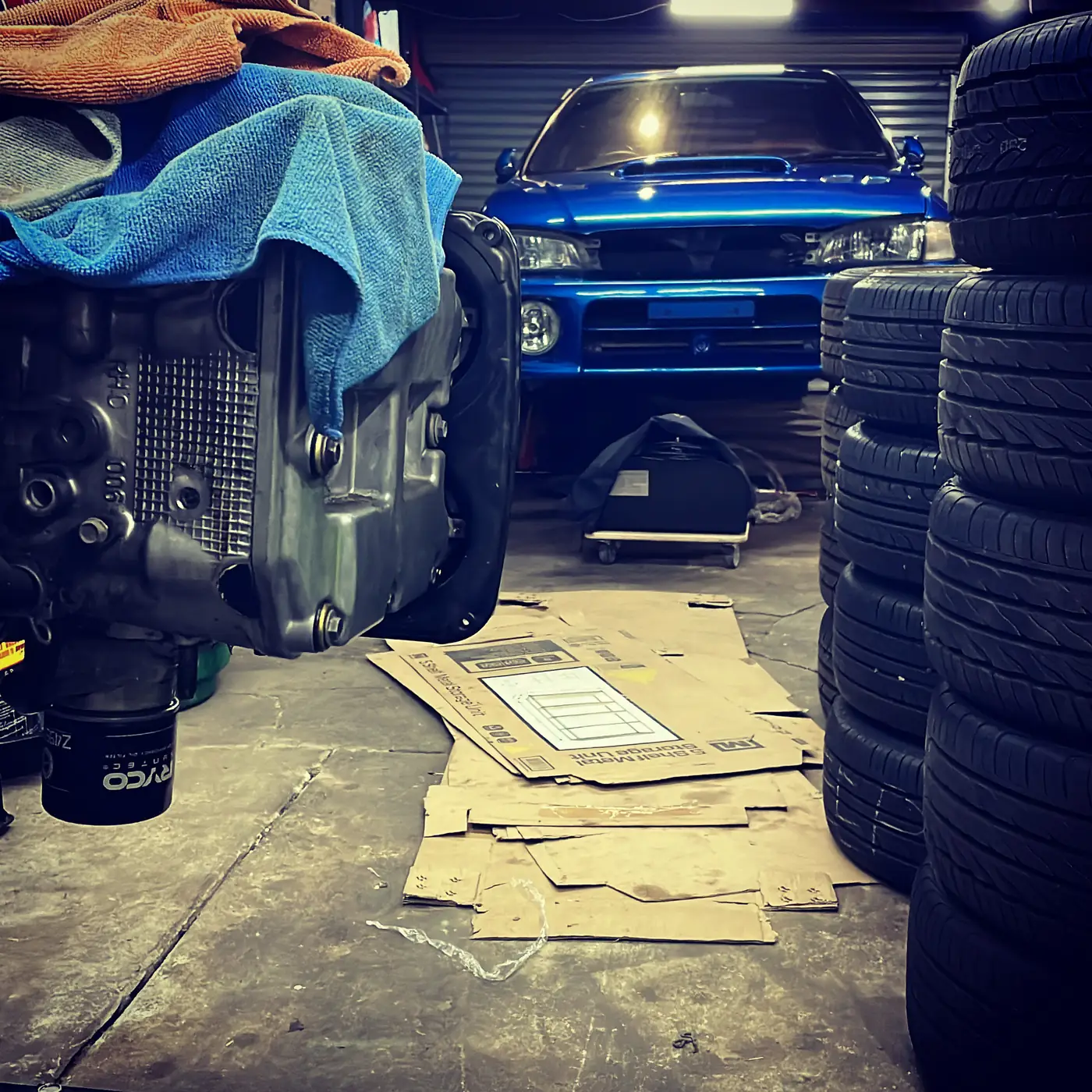 A garage floor mid-job: an engine sump and oil filter in the foreground under a draped microfibre cloth, flattened cardboard laid over the concrete, stacked tyres to one side, and the blue Impreza parked nose-on under a roller door.