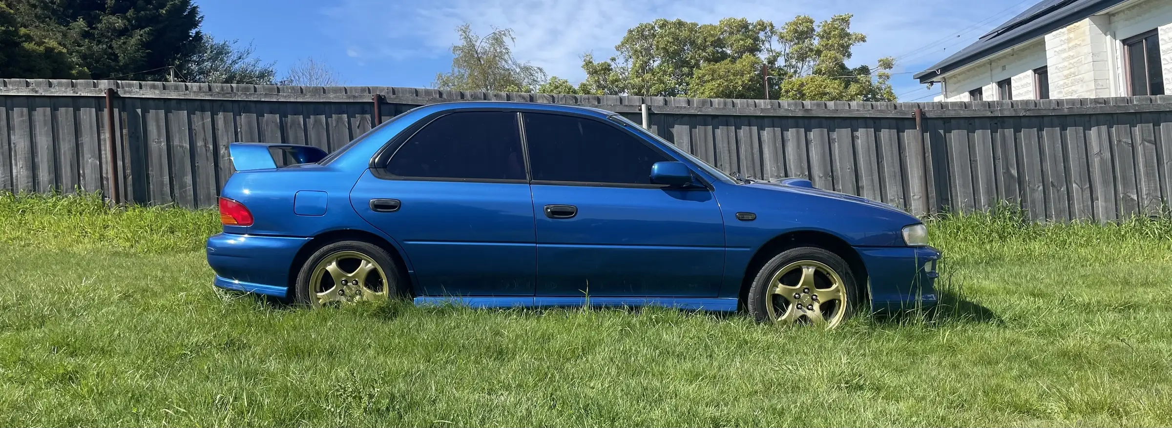 A 1998 Subaru Impreza WRX GC8 in blue with gold wheels, parked side-on against a corrugated fence.